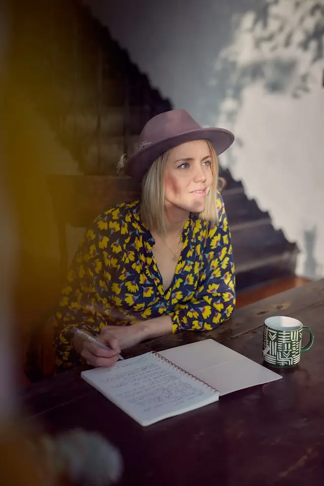 A woman in a patterned blouse and wide-brimmed hat writes in a notebook at a wooden table with a coffee mug beside her, looking thoughtfully out the window.
