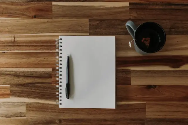 A top-down view of a wooden table with a spiral-bound notebook, a black pen placed vertically on the notebook, and a dark ceramic mug filled with tea. A tea bag string hangs over the rim of the mug. The scene suggests a workspace or a moment of quiet planning or reflection.